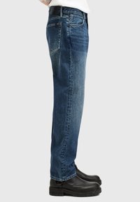 Jeans in denim blu con taglio dritto, caratterizzati da aree sbiadite, cinque tasche e una cintura nera. Abbinati a stivaletti neri alla caviglia.