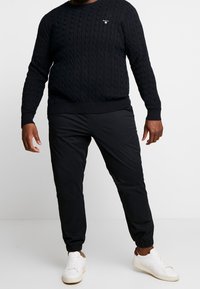 Marineblauwe kabelgebreide trui met geribbelde manchetten en onderrand, gecombineerd met zwarte joggers met elastische taille en witte sneakers.