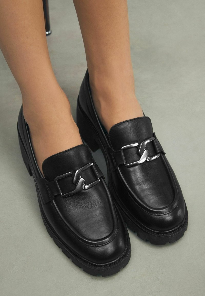 Mocassins en cuir noir avec détail chaîne argentée portés pieds nus, debout sur un sol lisse gris.