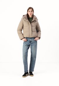 Beige péřová bunda s kapucí lemovanou kožešinou, na zip, s prošívaným vzorem, v kombinaci s modrými džínovými kalhotami a černými teniskami.