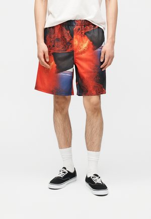 Homme portant un short multicolore à motifs abstraits rouges et noirs, des chaussettes blanches, des baskets noires et un t-shirt blanc uni, debout devant un fond neutre.