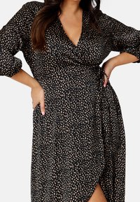 Schwarzes Wickelkleid mit beigefarbenem Punktmuster, tiefem V-Ausschnitt, Puffärmeln und einem seitlichen Schlitz. Hergestellt aus leichtem, strukturiertem Stoff.