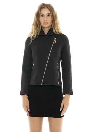 Une femme présente une veste noire à fermeture éclair asymétrique et une jupe noire ajustée sur fond blanc.