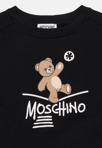 Μαύρο μπλουζάκι Moschino με γραφικό ενός καφέ αρκούδου που χαμογελά πάνω από το λογότυπο Moschino σε λευκό στυλιζαρισμένο κείμενο.