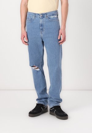 Mann trägt hellblaue, zerrissene Jeans mit geradem Bein und schwarze Turnschuhe, steht vor einem schlichten weißen Hintergrund.