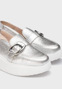 Zapatos plateados metálicos tipo slip-on con una superficie texturizada, que presentan un gran detalle de hebilla y una gruesa suela de plataforma blanca.
