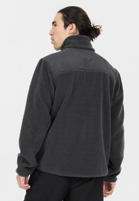 Tummanharmaa fleece-takki, jossa on korkea kaulus, teksturoitu kangas ja kiinteät harmaat olkapaneelit. Ominaisuuksiin kuuluvat pitkät hihat ja rento istuvuus.