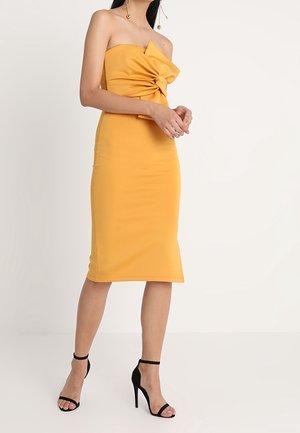 Robe moulante jaune sans bretelles avec un détail nœud à l'avant. Confectionnée dans un tissu lisse, de longueur mi-mollet, associée à des talons noirs à lanières.