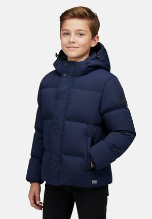 Jongen met kort bruin haar die een marineblauwe gewatteerde winterjas met capuchon en een zwarte broek draagt, staat met één hand in de zak.