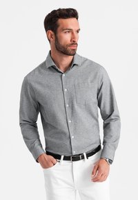 Chemise grise à boutons avec un motif texturé, dotée d'une poche poitrine, associée à un pantalon blanc et une ceinture noire, présentée sur un fond uni.