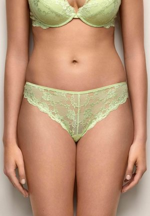 Donna che indossa un reggiseno in pizzo verde chiaro e slip coordinati in pizzo, in piedi contro uno sfondo semplice.