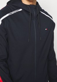 Marineblauer Zip-Hoodie mit weißen und roten Akzenten, ausgestattet mit einer Kapuze mit Zugband und einem Logo auf der Brust. Weicher Stoff mit glatter Textur.