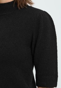 Sort strikket sweater med høj krave, korte pufærmer og ribbede manchetter. Teksturen virker blød og hyggelig, med en let fuzzy finish.