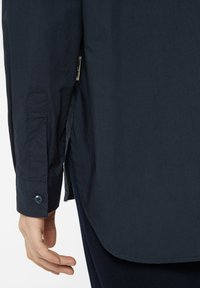 Navy long-sleeve shirt featuring a button cuff, slight curve at the hem, and a side vent with a fabric tag visible. Smooth texture.