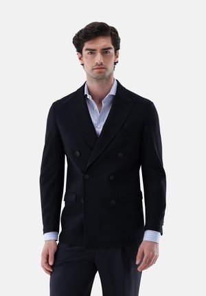 Mann, der einen dunklen zweireihigen Blazer über einem hellblau gestreiften Hemd trägt und vor einem weißen Hintergrund steht.