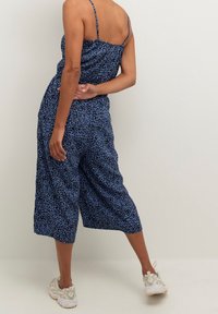 Femme portant une combinaison large à imprimé léopard bleu et des baskets blanches, posée avec un bras derrière le dos contre un fond uni.