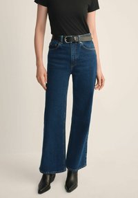 Kobieta w ciemnoniebieskich, szerokich spodniach z wysokim stanem, czarnym pasku, czarnym topie z krótkim rękawem oraz czarnych, szpiczastych botkach na kostkę na beżowym tle.