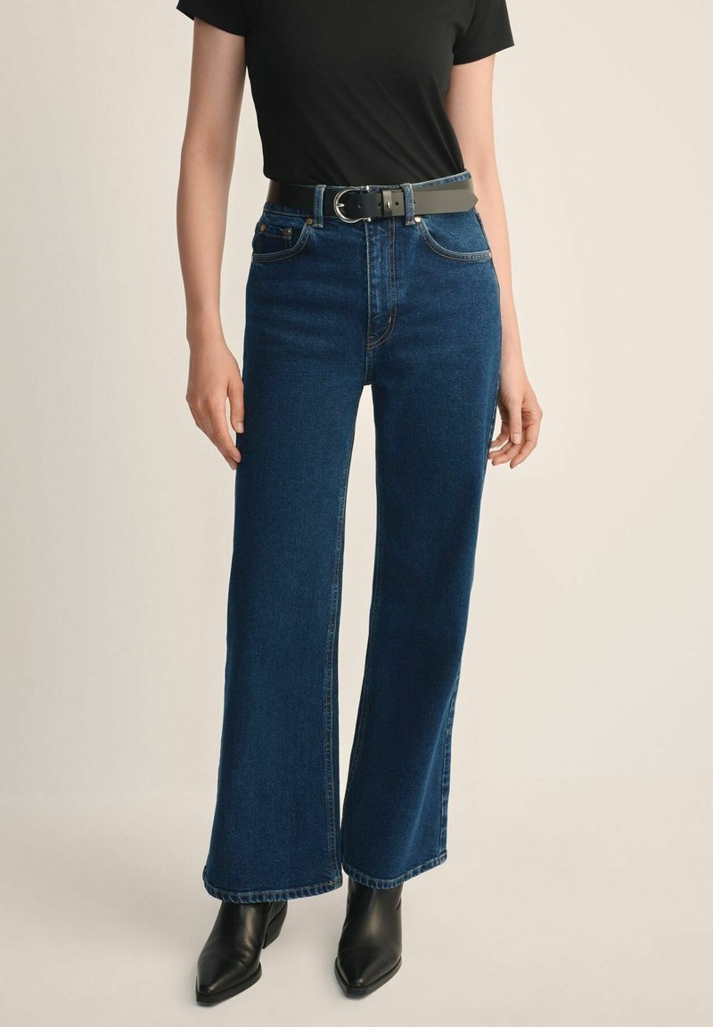 Kobieta w ciemnoniebieskich, szerokich spodniach z wysokim stanem, czarnym pasku, czarnym topie z krótkim rękawem oraz czarnych, szpiczastych botkach na kostkę na beżowym tle.