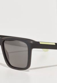 Ochelari de soare rectangulari negri cu lentile gri, având un finisaj mat și un accent verde pe întreruperi. Logo-ul "Polaroid" pe ramă.