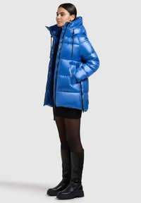 Pufferjacka i klarblå färg med huva, dragkedja och sidorfickor med svarta detaljer. Mjuk, glänsande textur med quiltat mönster.