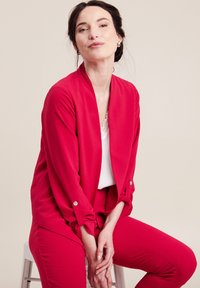 Giacca leggera rossa con apertura drappeggiata e maniche lunghe. Abbinata a pantaloni rossi coordinati, con una texture liscia e una vestibilità sartoriale.