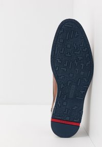 Chaussure en cuir marron avec une semelle en caoutchouc bleu texturé, présentant des motifs géométriques et une bande d'accent rouge au niveau du talon.