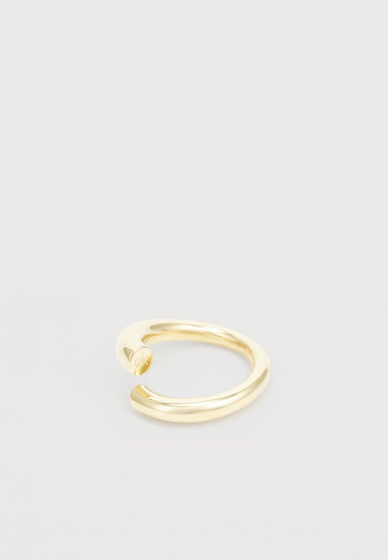 Anillo en tono dorado con un diseño suave y redondeado, con una apertura en un extremo y detalle de logo grabado. Hecho de metal pulido.
