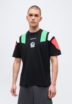 Bărbat purtând un tricou negru Adidas cu dungi verzi, albe și roșii pe umeri și emblema de fotbal a Mexicului, stând în fața unui fundal alb.