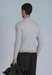 Hellgrauer, gerippter Pullover mit langen Ärmeln, taillierten Schnitt, Rundhalsausschnitt und einem Saum, der zu einer dunklen Hose passt, die mit einer schwarzen Jacke getragen wird.