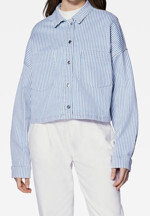 Kobieta ubrana w krótki, niebiesko-biały koszulowy top w pionowe paski z dwoma kieszeniami na piersi oraz białe spodnie z wysokim stanem.