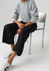 Pull gris clair en maille avec poignets côtelés, pantalon large noir, et baskets blanches avec accents noirs, assis sur une chaise en métal.