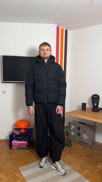 Juoda pūkinė striukė su užtrauktuku priekyje ir kapišonu; turi siuvinėtus skersinius. Dėvima su laisvomis juodomis kelnėmis ir pilkais sportbačiais.