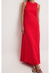 Femme portant une robe rouge sans manches, longue jusqu'au sol, en tissu texturé, debout devant un fond uni de couleur claire.