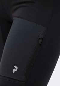 Κοντινό πλάνο μαύρων αθλητικών κολάν με φερμουάρ στην τσέπη στο μηρό και αντανακλαστικό λογότυπο στο κάτω μέρος του μηρού.