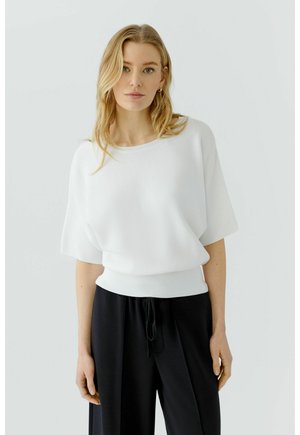 Blonde Frau mit welligem Haar trägt ein weißes Kurzarm-Top und weite schwarze Hosen und steht vor einem schlichten, hellen Hintergrund.