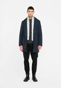 Cappotto lungo blu navy con bottoni frontali, gilet grigio sotto, maglione lavorato a maglia grigio e pantaloni neri. Scarpe nere alla caviglia completano l'outfit.