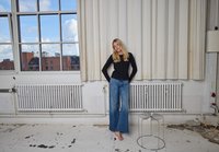 Cardigan nero con bottoni e jeans blu a gamba larga; radiatore bianco e sgabello in metallo; tende leggere; grande finestra con vista sulla città.