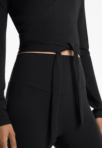 Black long-sleeve top with a wrap design and tied detail at the waist, paired with high-waisted black leggings. Smooth texture, fitted shape.
