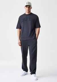 Siva oversized majica s povišenim napisom "NEW ERA", v kombinaciji s ujemajočimi se ozkimi trenirkami in belimi supergami, oblečeno s kapo.