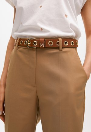 Ce ceinturon en faux cuir marron avec des accessoires en or présente des œillets circulaires et un logo bien en vue. Il est associé à un pantalon beige.
