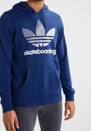 Mężczyzna w niebieskiej bluzie z kapturem z białym paskowanym logo koniczyny i napisem „skateboarding”, w połączeniu z ciemnoszarymi jeansami.