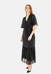 Vestido largo negro con mangas cortas acampanadas, escote en V profundo, textura acanalada, cintura ajustada con cinturón de lazo y detalle de dobladillo con volantes.