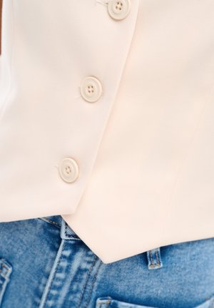 Cream-colored buttoned garment over blue denim jeans, showing three buttons and the waistband area.