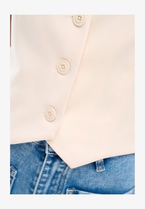 Cream-colored buttoned garment over blue denim jeans, showing three buttons and the waistband area.