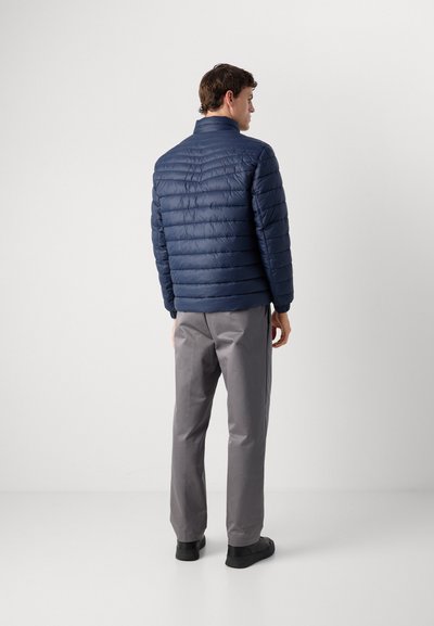 Homme de dos portant une veste matelassée bleu marine, un pantalon gris et des chaussures noires sur un fond blanc uni.