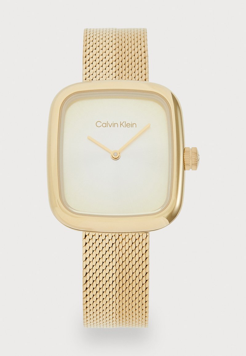 Auksinė kvadratinė laikrodžio su tinkline apyranke, turi šviesios spalvos ciferblatą ir paprastas valandų bei minučių rodykles. Viršuje yra prekės ženklo pavadinimas Calvin Klein.