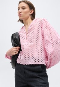 Naine, kes kannab heleroosat õhulise pitsist pluusi puhvvarrukatega, musti pükse, hoides käes korralikult voltitud musta nahkjopet, neutraalse taustaga.