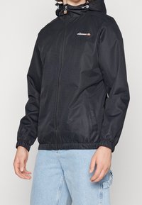 Czarna kurtka z kapturem, zapinana na suwak z przodu, z elastycznymi mankietami i dołem. Na lewej piersi znajduje się logo marki, a materiał ma teksturowane wykończenie.