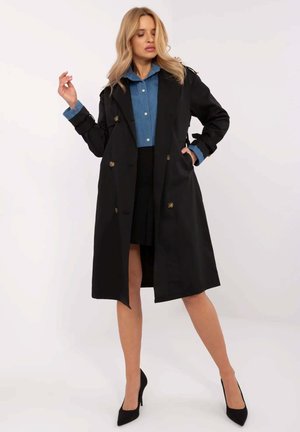 Blondynka ubrana w czarny płaszcz trencz, niebieską koszulę zapinaną na guziki, czarną spódnicę i czarne wysokie obcasy stoi z jedną ręką w kieszeni.