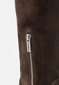 Bota de ante marrón hasta el tobillo con una cremallera plateada en el lateral, con una etiqueta de marca visible que dice "SHABBIES". Textura suave y diseño elegante.